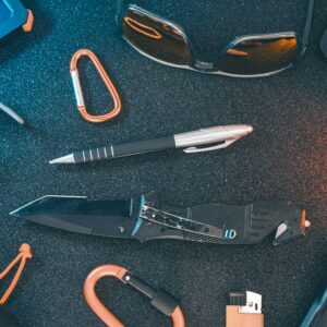 A flat lay of essential everyday carry items including a knife, pen, sunglasses, and carabiner.