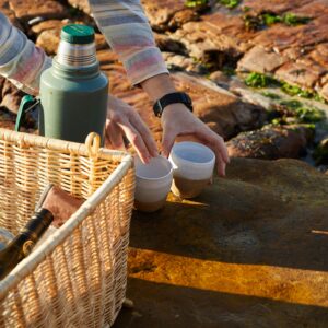 Preparing for a seaside picnic with a wicker basket, cups, and a thermos on rocky terrain.