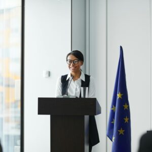 A woman giving a speech at a podium with an EU flag in a conference setting.