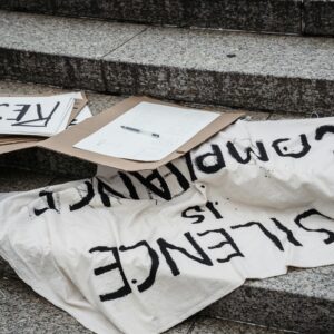 A close-up of protest signs reading 'RACIST' and 'Silence is Complacency' displayed on stone steps.
